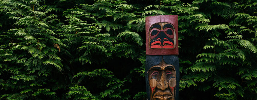 A carved Indigenous pole stands prominently in front of a lush green forest backdrop in Kia'palano, showcasing the rich cultural heritage and artistic traditions preserved at Capilano Suspension Bridge Park.