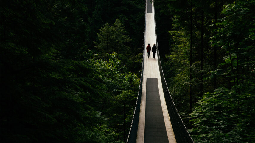 A silhouette of two guests walking in the middle of the iconic Capilano Suspension Bridge, surrounded by breathtaking natural beauty