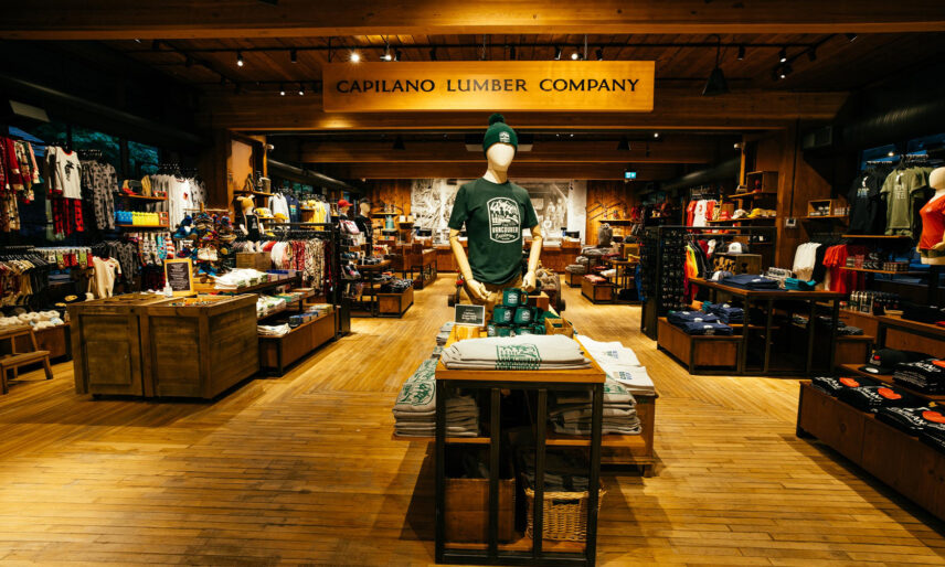 An area of the Trading Post Gift Shop features various clothing items and a large wooden banner reading 'Capilano Lumber Company,' blending modern apparel with nods to the park's historical logging heritage at Capilano Suspension Bridge Park.