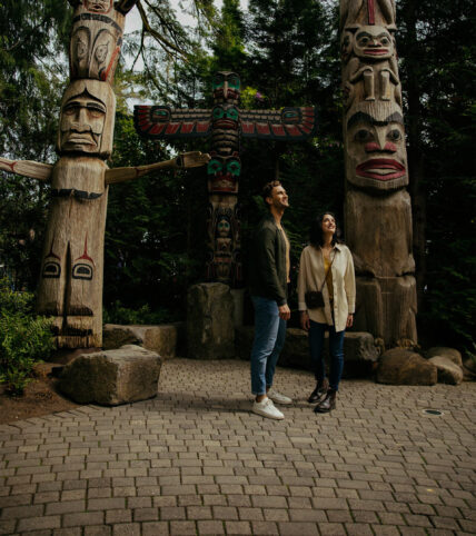Two guests stand surrounded by towering totem poles in Kia'palano, admiring the rich cultural heritage and artistic craftsmanship at Capilano Suspension Bridge Park
