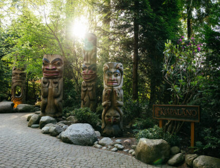 The entrance sign of Kia'palano at Capilano Suspension Bridge Park, framed by majestic totem poles with the morning sun peeking through, welcoming visitors to an immersive journey into Pacific Northwest culture and heritage
