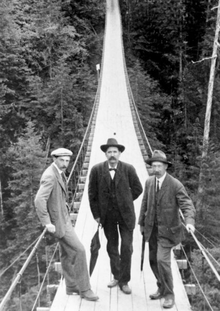 An old black and white photograph from 1913 featuring three men standing proudly on the Capilano Suspension Bridge, offering a glimpse into its rich history and legacy as a marvel of engineering and adventure