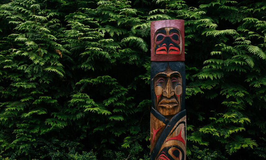 A carved Indigenous pole stands prominently in front of a lush green forest backdrop in Kia'palano, showcasing the rich cultural heritage and artistic traditions preserved at Capilano Suspension Bridge Park.