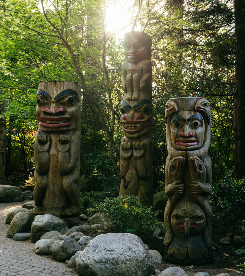 The entrance sign of Kia'palano at Capilano Suspension Bridge Park, framed by majestic totem poles with the morning sun peeking through, welcoming visitors to an immersive journey into Pacific Northwest culture and heritage