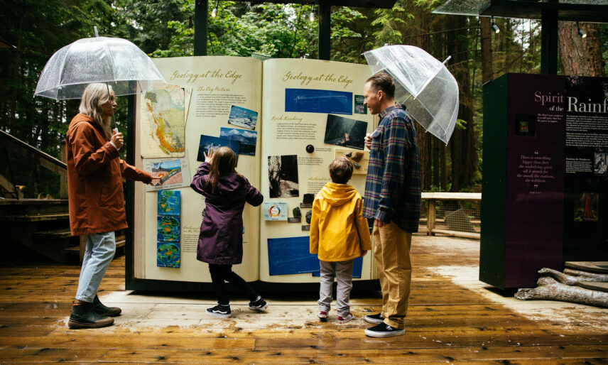On a rainy day, a family interacts with the Living Forest exhibit panels. The parents hold umbrellas while the kids eagerly examine the displays, engaged and curious about the wonders of the rainforest at Capilano Suspension Bridge Park.