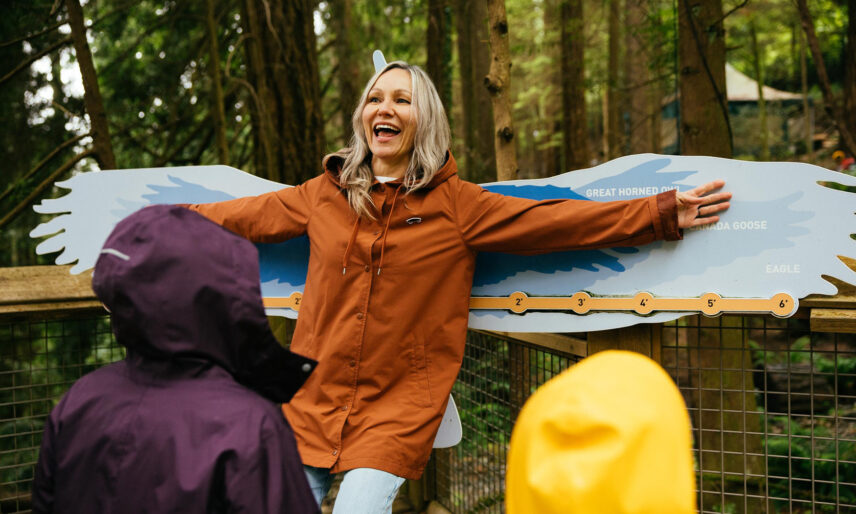 A mother extends her arms out to a bird wing display while her two kids watch eagerly in the foreground, captivated by the interactive Living Forest exhibits at Capilano Suspension Bridge Park