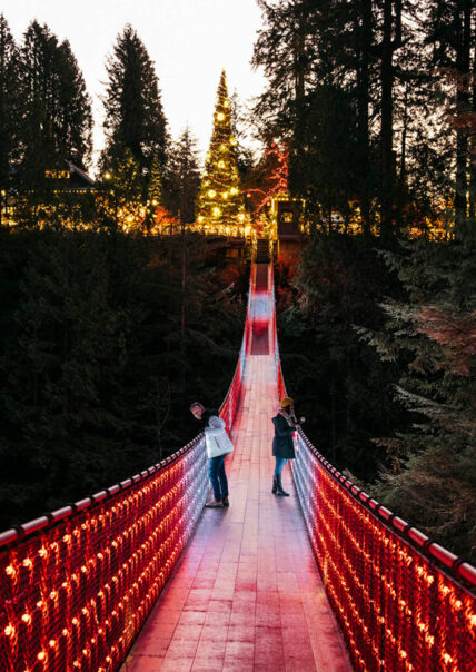 A couple looking out from the Capilano Suspension Bridge illuminated in vibrant red and white hues during the Love Lights event, creating a romantic atmosphere against the evening backdrop