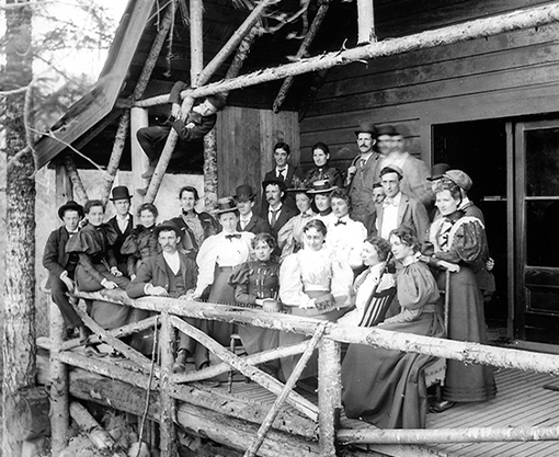 A historical black and white photo depicts a group of settlers known as the Capilano Tramps visiting the Park back when the bridge was first built, highlighting the bridge's early days and its role in attracting visitors to the area.