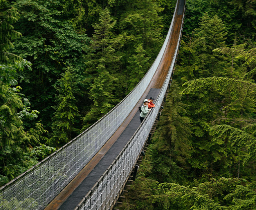 Two visitors enjoy a thrilling stroll across the iconic Capilano Suspension Bridge, suspended high above the lush greenery of the rainforest below