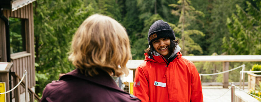 A friendly team member of Capilano Suspension Bridge Park warmly smiles and greets a guest approaching them, embodying the welcoming atmosphere and hospitality of the park.