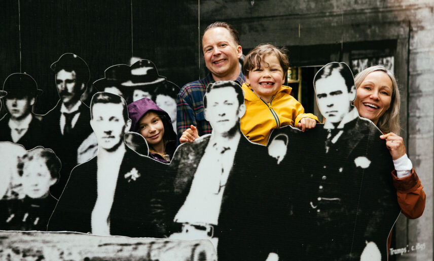 A family smiles and poses in front of an interactive display featuring a black and white photo of the Capilano Tramps, engaging with the rich history and heritage of Capilano Suspension Bridge Park.