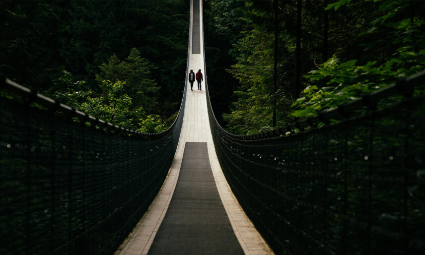 Two guests enjoy a leisurely stroll on the Capilano Suspension Bridge, set against the backdrop of lush greenery and towering trees, experiencing the awe-inspiring beauty of Capilano Suspension Bridge Park