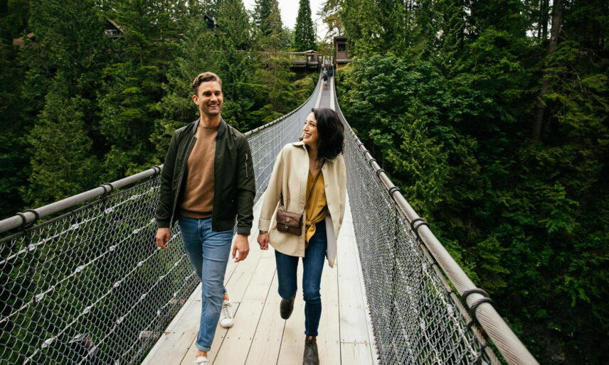 A couple enjoys a romantic stroll on the Capilano Suspension Bridge, smiling at each other as they embrace the adventure and scenic beauty of Capilano Suspension Bridge Park.