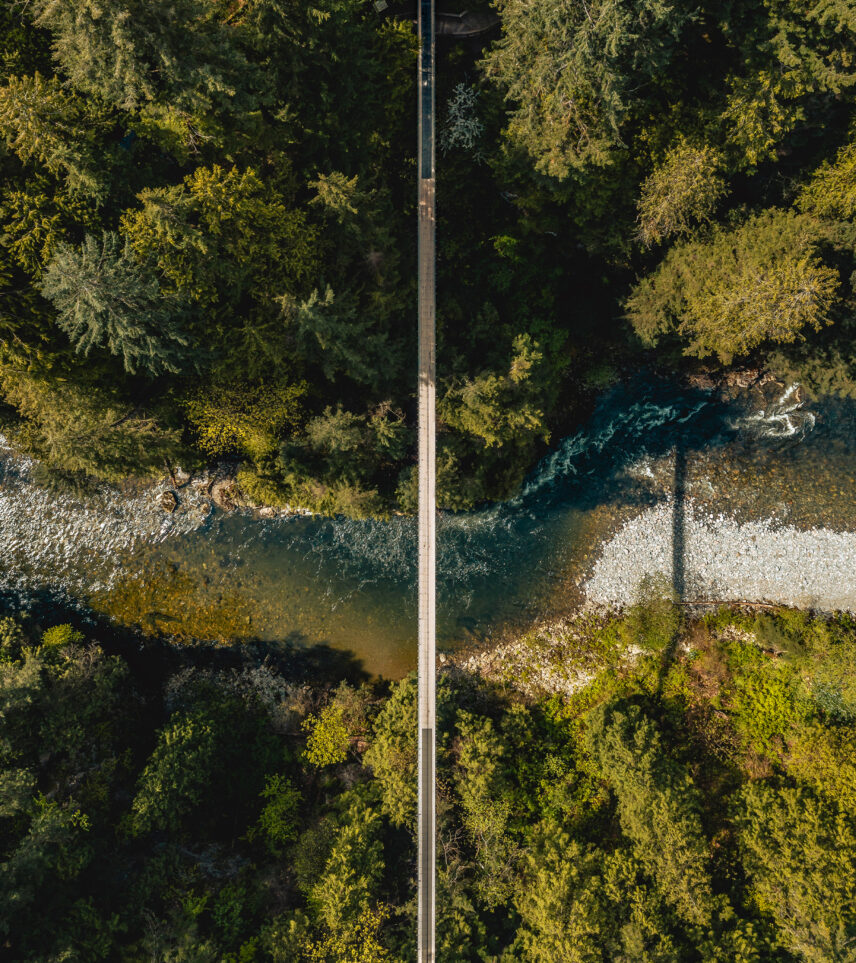An aerial view directly above the Capilano Suspension Bridge, showcasing the lush rainforest and winding river below, offering a stunning perspective of this iconic landmark in its natural setting