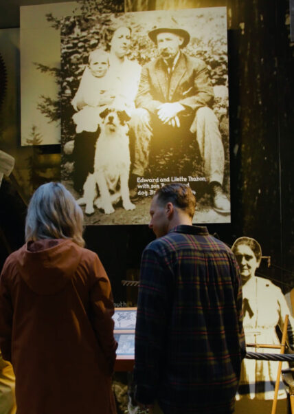 A family gathers around a historical panel in the Story Centre at Capilano Suspension Bridge Park, eagerly reading about the rich heritage and captivating tales associated with the park's iconic landmark