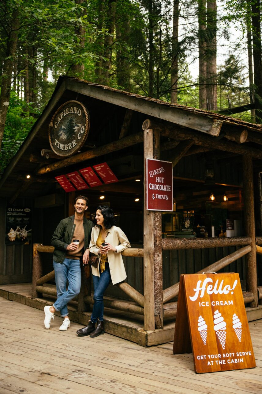 two guests standing in front of the Cabin cafe drinking coffee surrounded by a green forest at Capilano Suspension Bridge Park