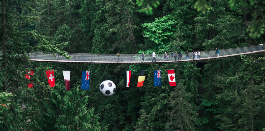 the capilano suspension bridge with 8 country flags hanging underneath it and a large soccer ball hangs in the middle for the Canyon Kick-Off event.