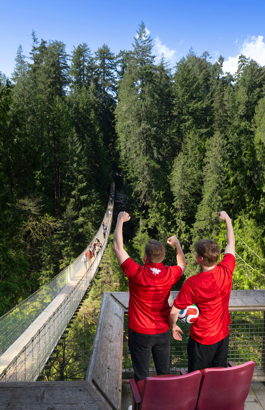 Two Canada soccer fans standing and cheering on a deck at Capilano Suspension Bridge Park during the Canyon Kick-Off event, with stadium seats behind them and a rainforest canyon view in the background.